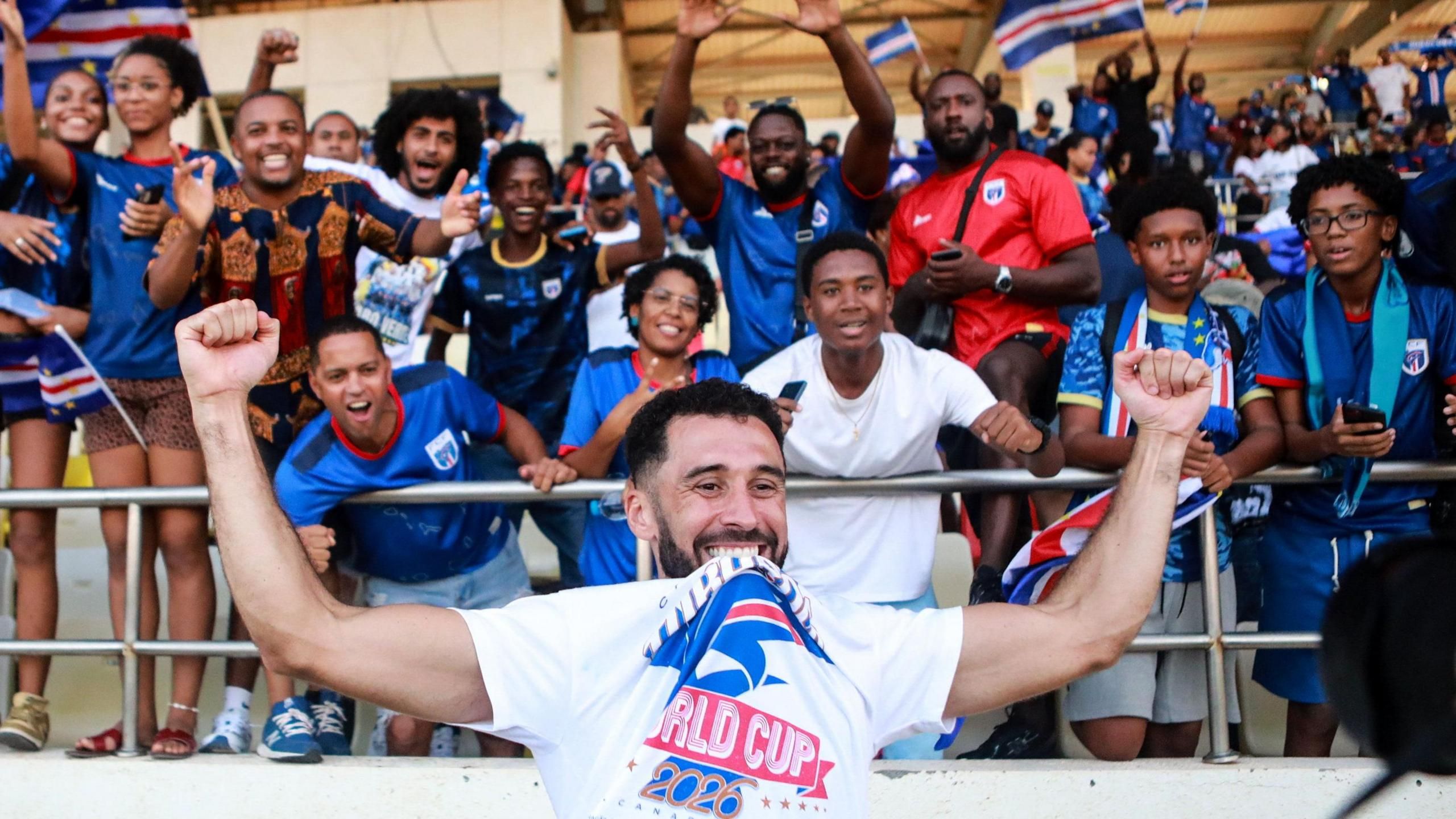Roberto Lopes، visible from chest up in the foreground of the image، holds his arms up high as he celebrates Cape Verde qualifying for the 2026 World Cup. He wears a white T-shirt with the text World Cup 2026 on it، which he raises up by biting it between his teeth. Twelve joyful Cape Verde fans، mostly wearing blue team jerseys، are seen in the stands behind him، with other people and some Cape Verde flags seen out of focus in the distance