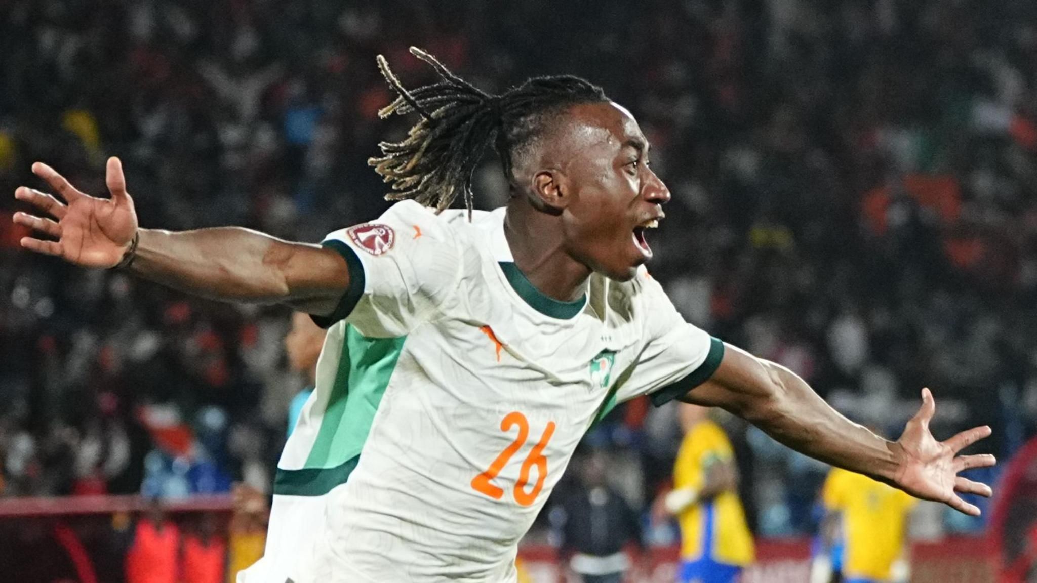 Yan Diomande، wearing a white Ivory Coast shirt with an orange number 26 on the chest، spreads his arms wide and opens his mouth as he wheels away in celebration، his hair seen flowing behind him. A packed crowd in a football stadium is out of focus in the background
